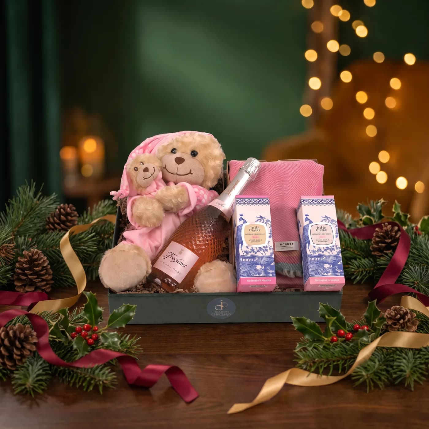 The Hello Baby Girl hamper dressed for Christmas, a soft bedtime bear with its comforter beside a bottle of Freixenet sparkling rosé, Julie Clarke hand wash and lotion and a pink lambswool blanket