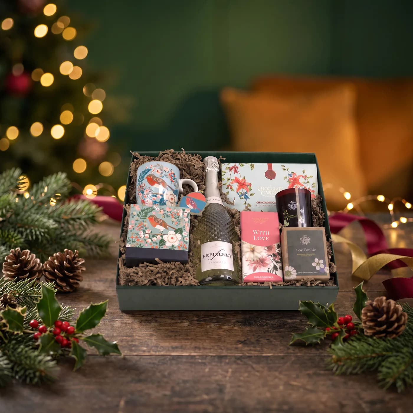 The Birdy Awesome hamper dressed for Christmas, a robin-patterned mug and notebook beside a bottle of Freixenet Prosecco, a boxed soy candle and chocolates, in a gift box on a wooden table
